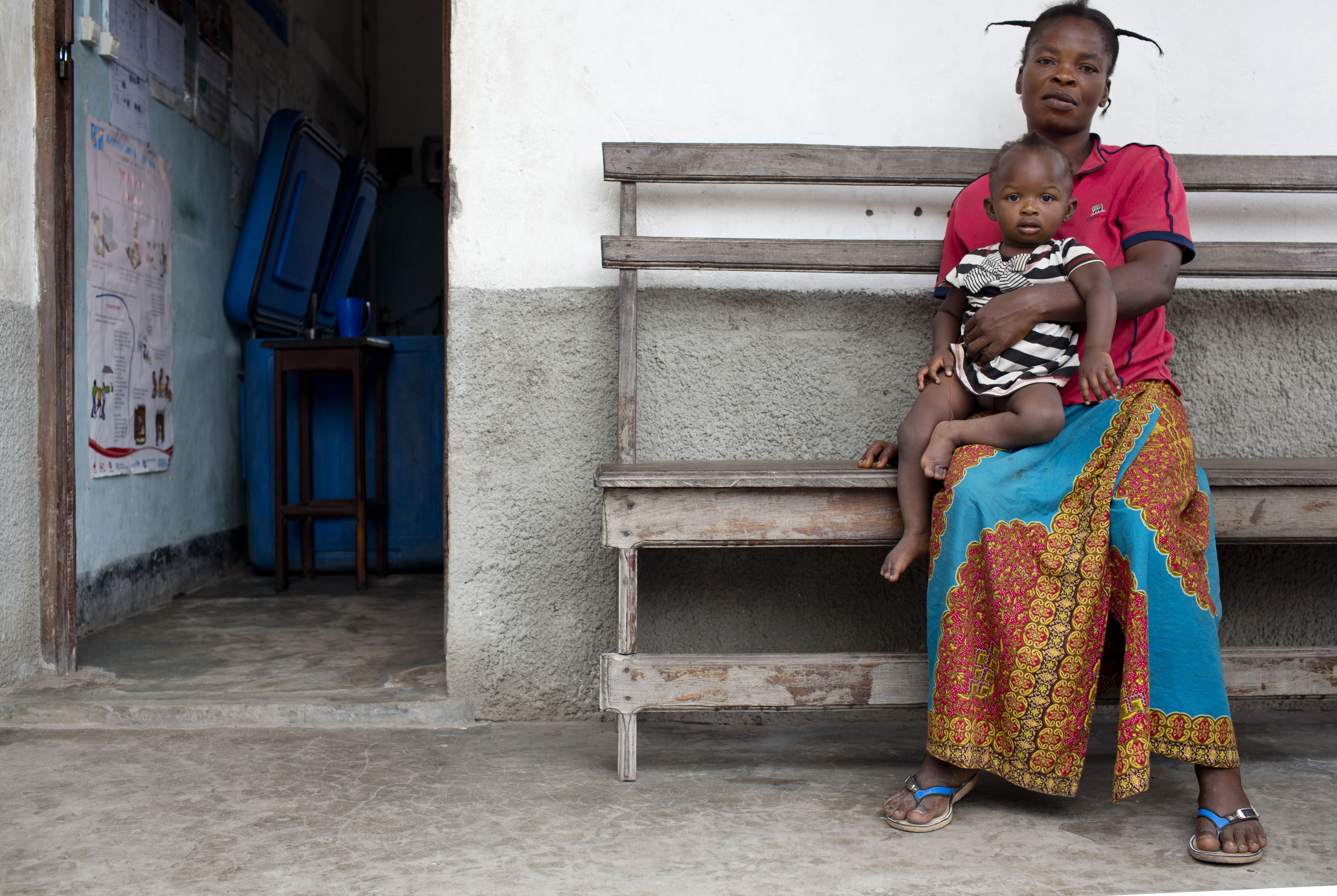 African mother sitting outside of a health post with a baby on her lap.